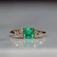 Close-cropped macro of a vintage ring, featuring a square emerald-cut emerald flanked by three round diamonds arranged in a triangular cluster on each shoulder. Viewed head-on.