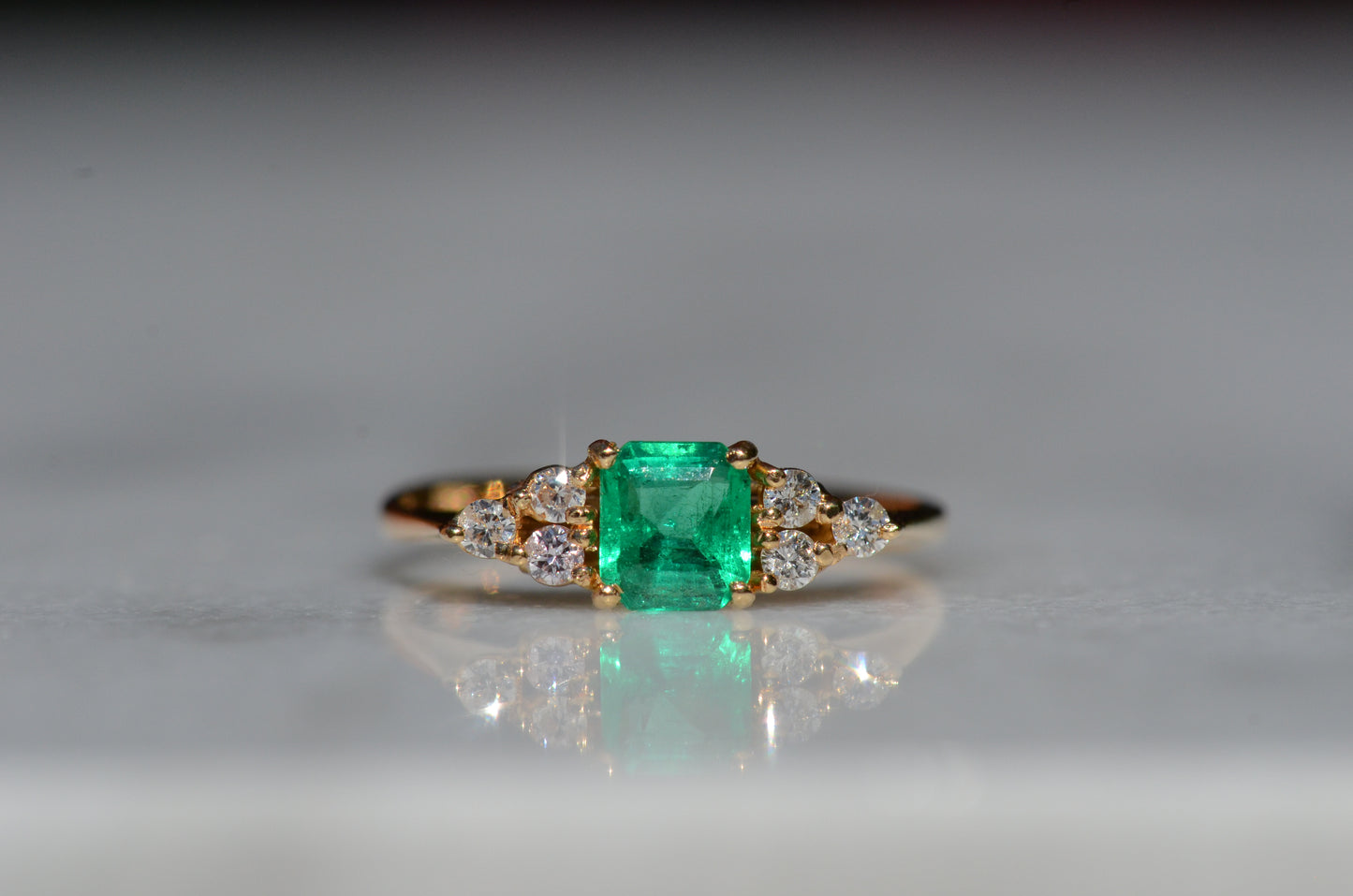 Close-cropped macro of a vintage ring, featuring a square emerald-cut emerald flanked by three round diamonds arranged in a triangular cluster on each shoulder. Viewed head-on.