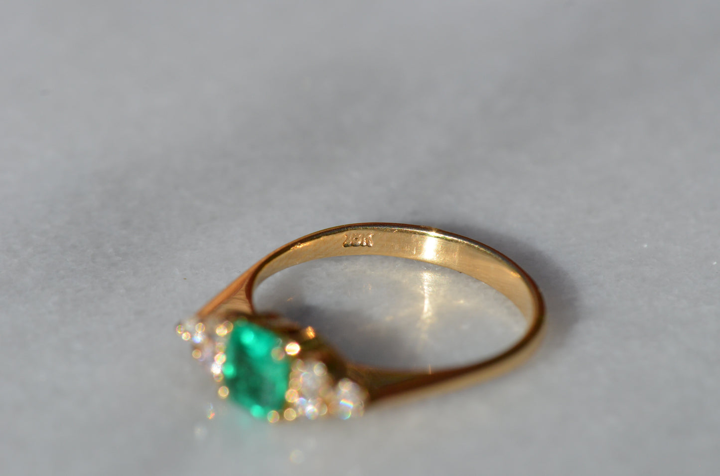 Close-cropped macro of a vintage ring, featuring a square emerald-cut emerald flanked by three round diamonds arranged in a triangular cluster on each shoulder. Viewed looking at the inside of the band to show the gold purity stamp.
