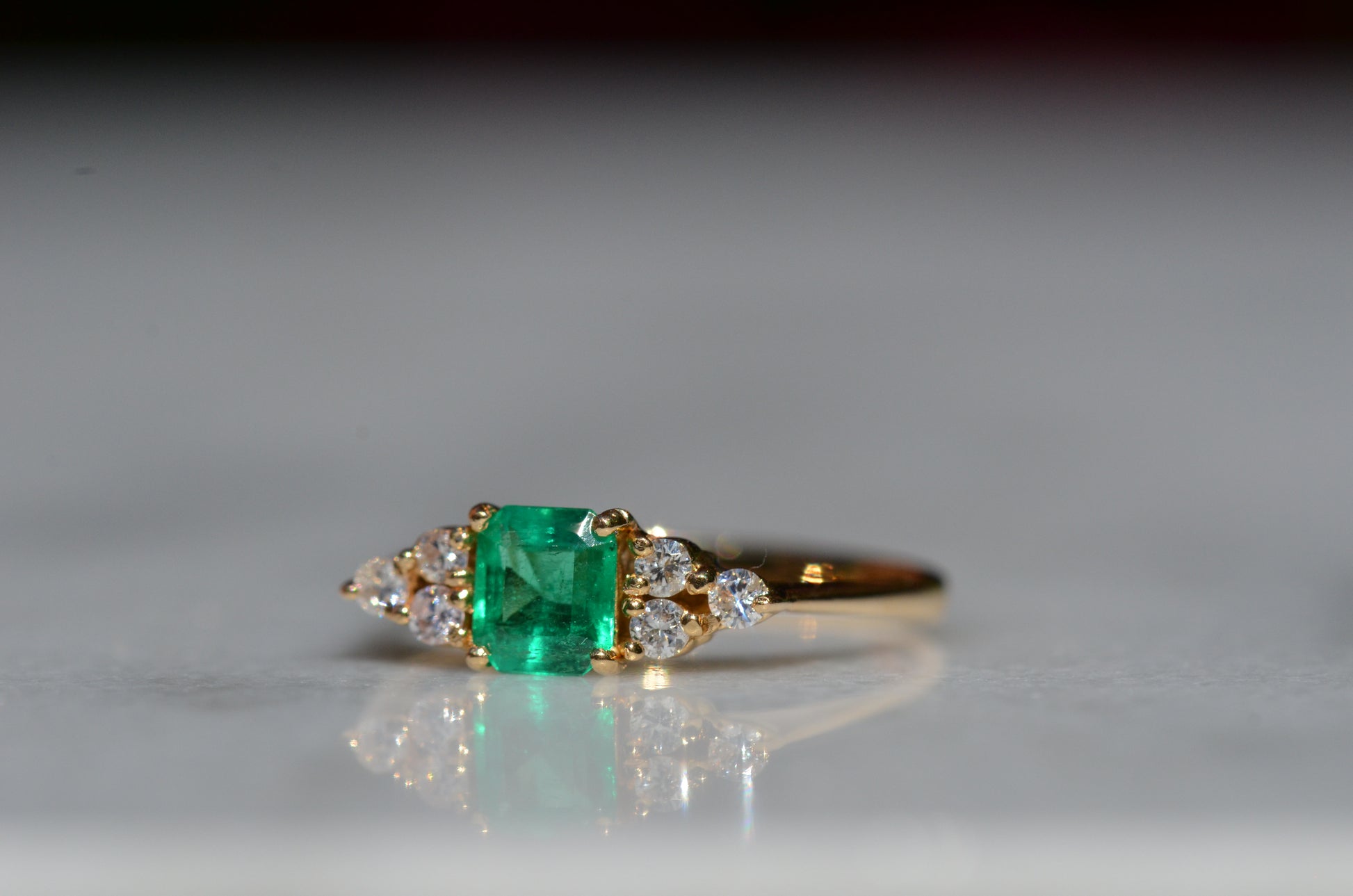 Close-cropped macro of a vintage ring, featuring a square emerald-cut emerald flanked by three round diamonds arranged in a triangular cluster on each shoulder. Viewed turned slightly to the left to highlight the shoulders.