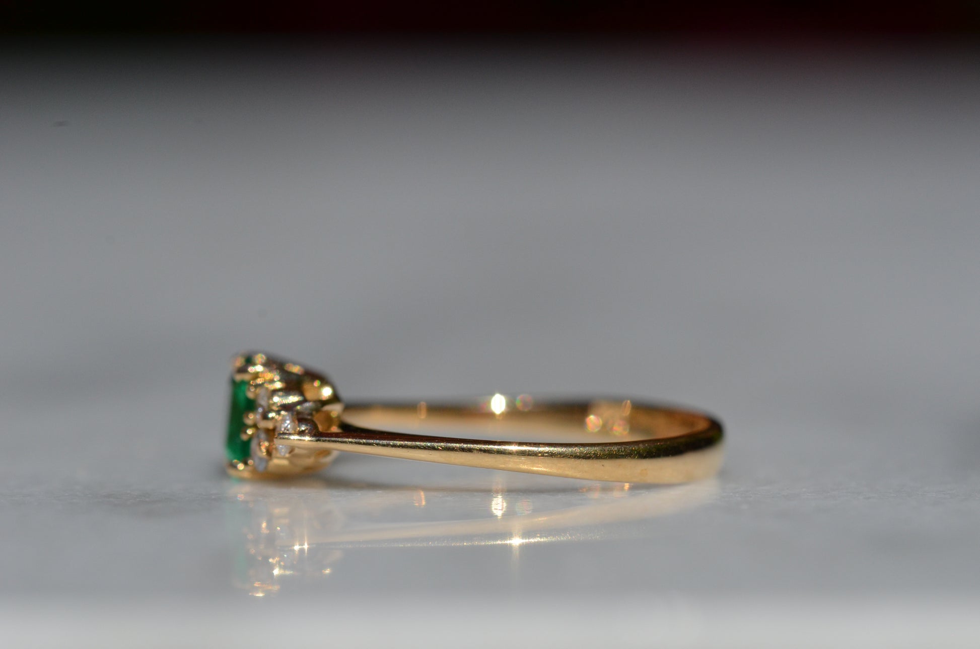 Close-cropped macro of a vintage ring, featuring a square emerald-cut emerald flanked by three round diamonds arranged in a triangular cluster on each shoulder. Viewed turned to the left to highlight the side of the band.