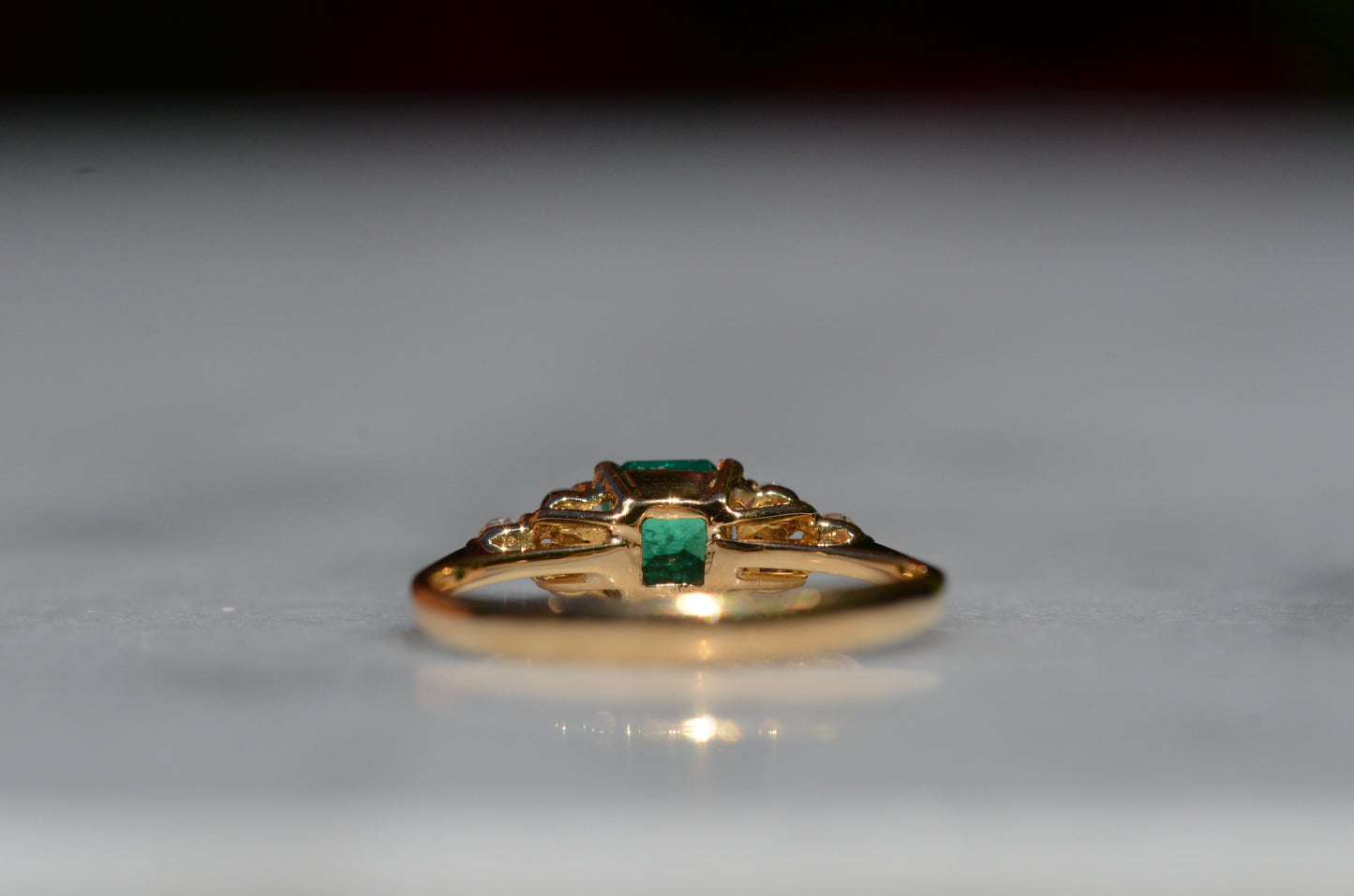 Close-cropped macro of a vintage ring, featuring a square emerald-cut emerald flanked by three round diamonds arranged in a triangular cluster on each shoulder. Viewed from the back focusing on the underside of the mount and back of the gemstones.
