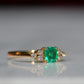 Close-cropped macro of a vintage ring, featuring a square emerald-cut emerald flanked by three round diamonds arranged in a triangular cluster on each shoulder. Viewed turned to the right to highlight the side of the ring.