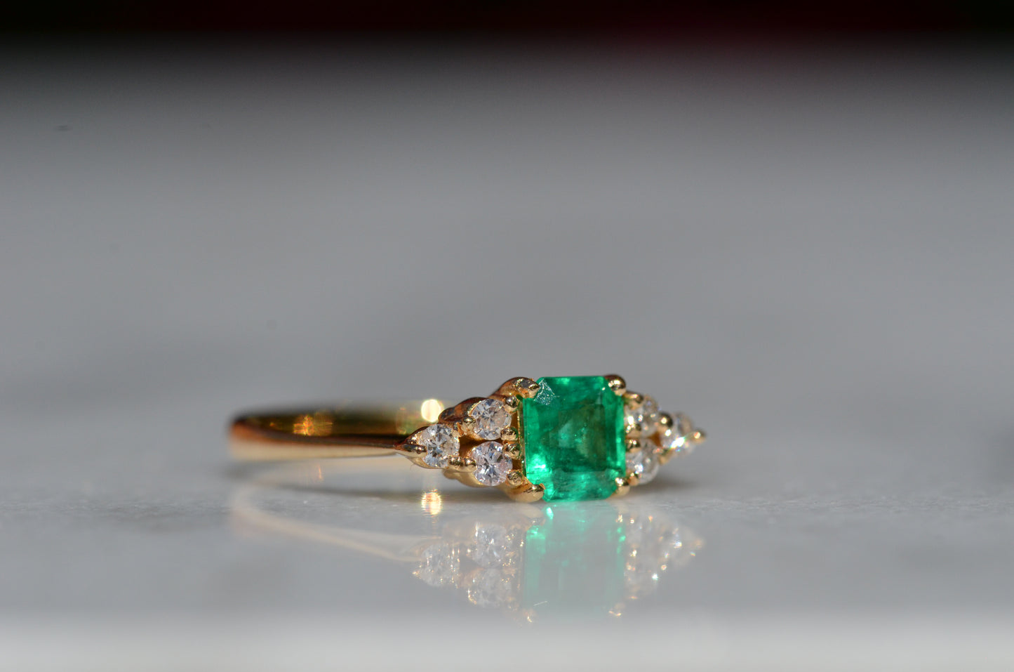 Close-cropped macro of a vintage ring, featuring a square emerald-cut emerald flanked by three round diamonds arranged in a triangular cluster on each shoulder. Viewed turned to the right to highlight the side of the ring.