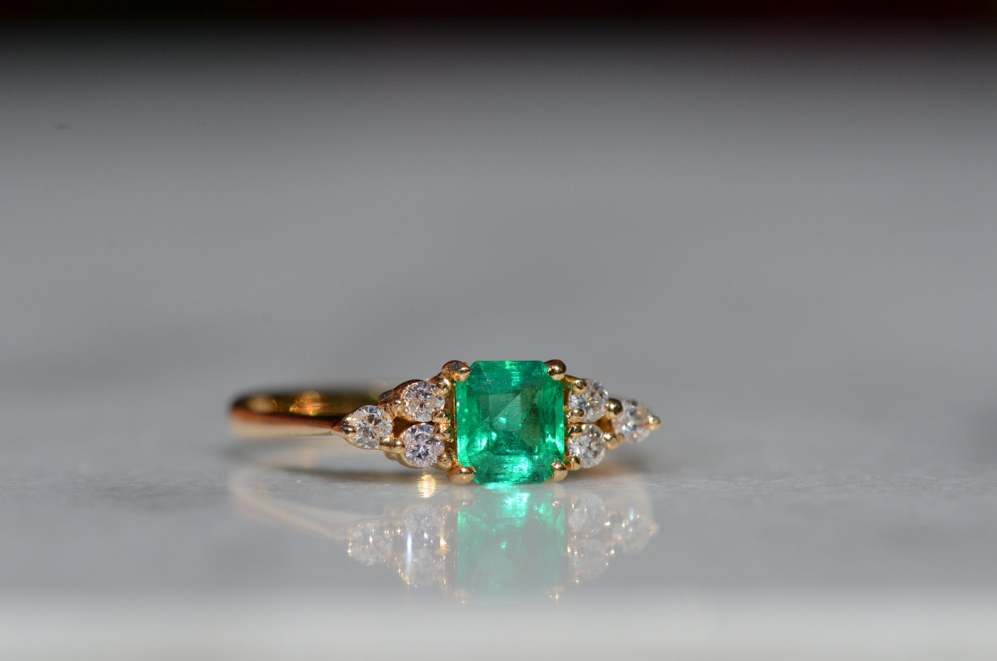 Close-cropped macro of a vintage ring, featuring a square emerald-cut emerald flanked by three round diamonds arranged in a triangular cluster on each shoulder. Viewed slightly to the right to highlight the diamond shoulders.