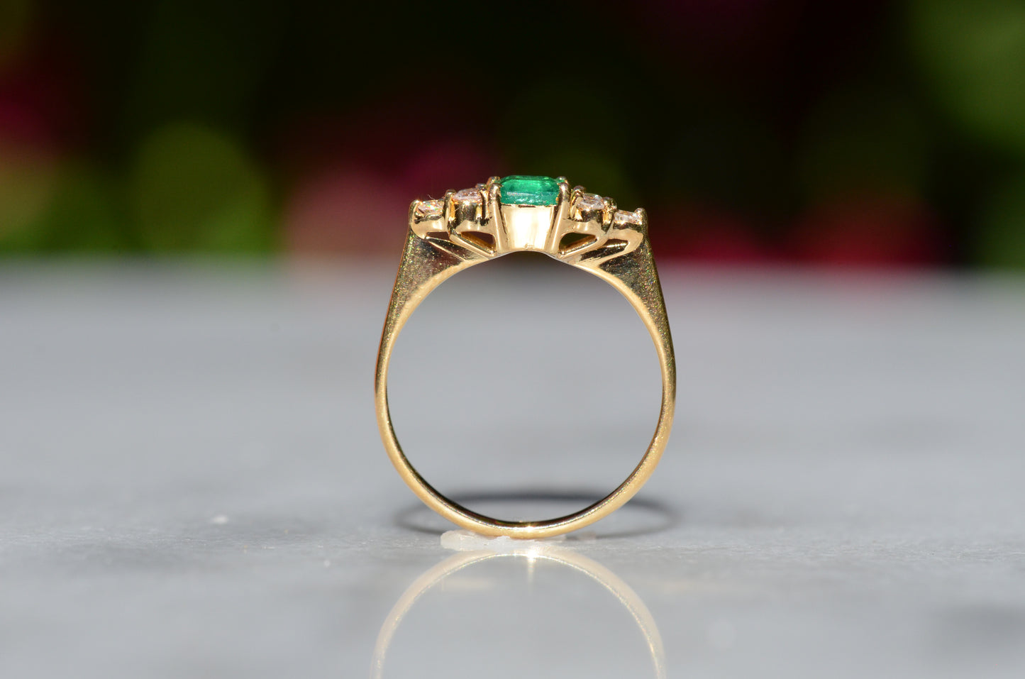 Close-cropped macro of a vintage ring, featuring a square emerald-cut emerald flanked by three round diamonds arranged in a triangular cluster on each shoulder. Viewed upright and looking through the finger hole to highlight the gallery of the mounting.